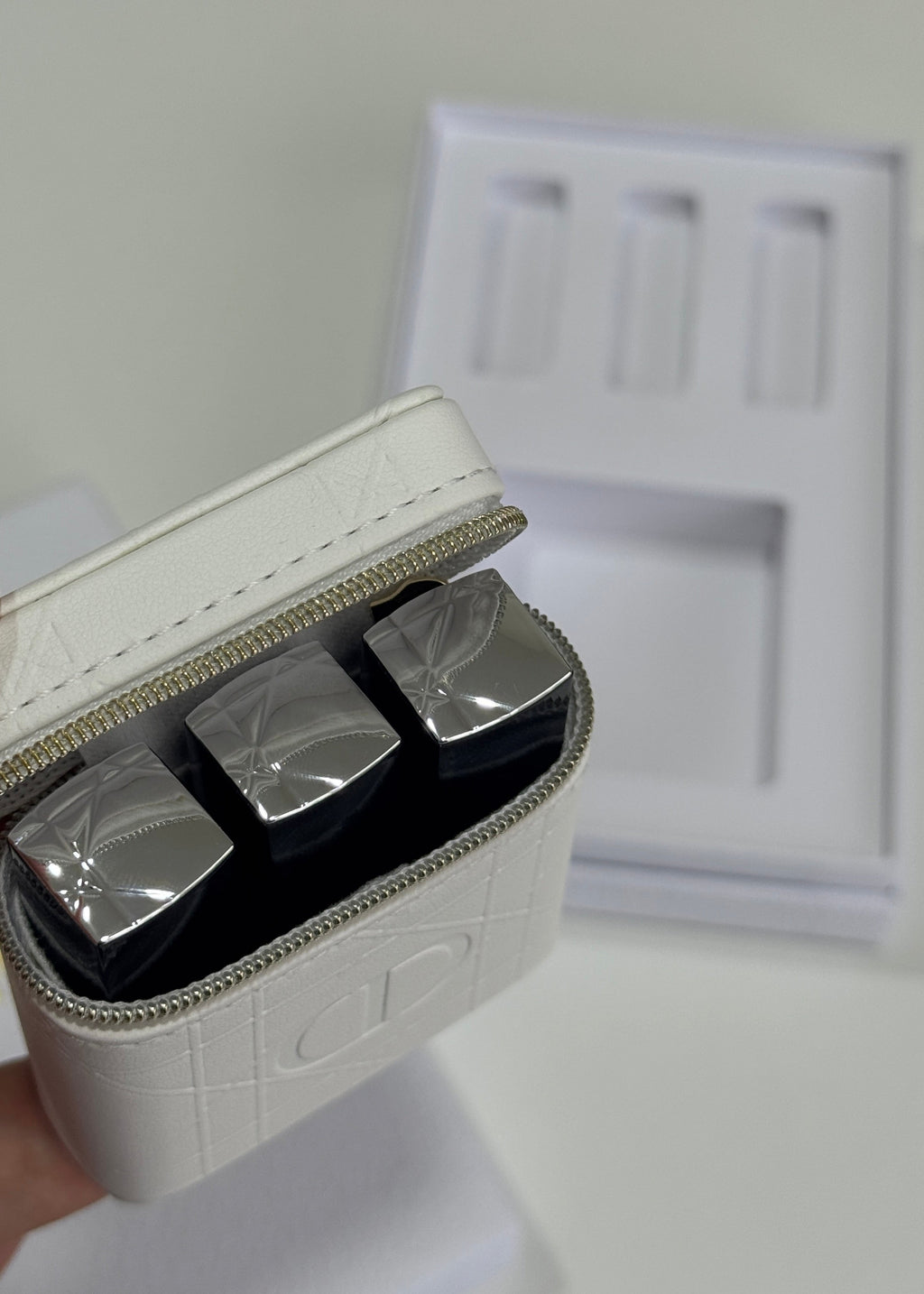 A close-up of a white textured zippered cosmetic case holding three silver rectangular items, with a white box insert and packaging in the background.