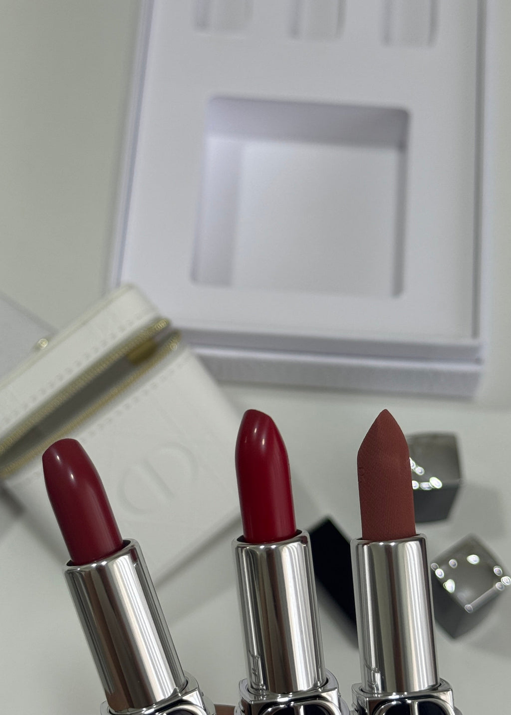 Three Dior lipstick tubes held in a hand, showing red, deep berry, and nude shades, with the white Dior box and silver cases in the background.
