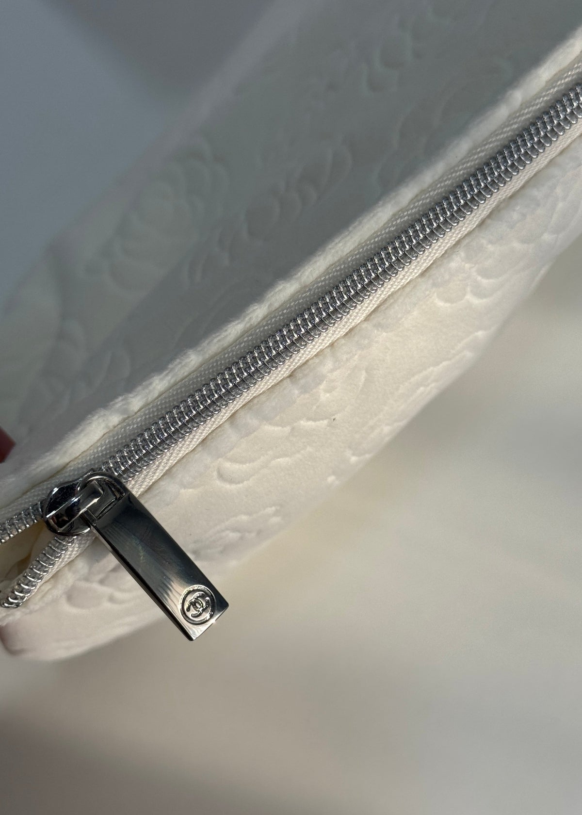 Close-up of a white embossed leather zippered pouch with a silver metal zipper and a black pull tab featuring a circular logo.