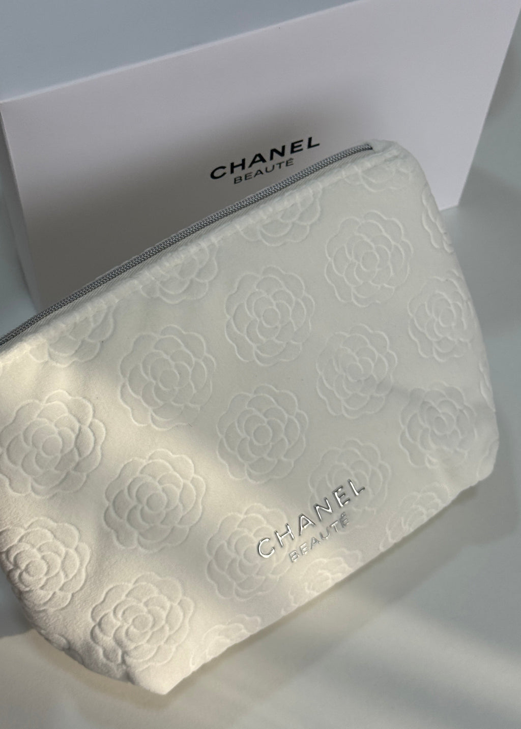 A white Chanel beauty pouch with embossed floral patterns and a silver zipper, shown in front of its white Chanel box.