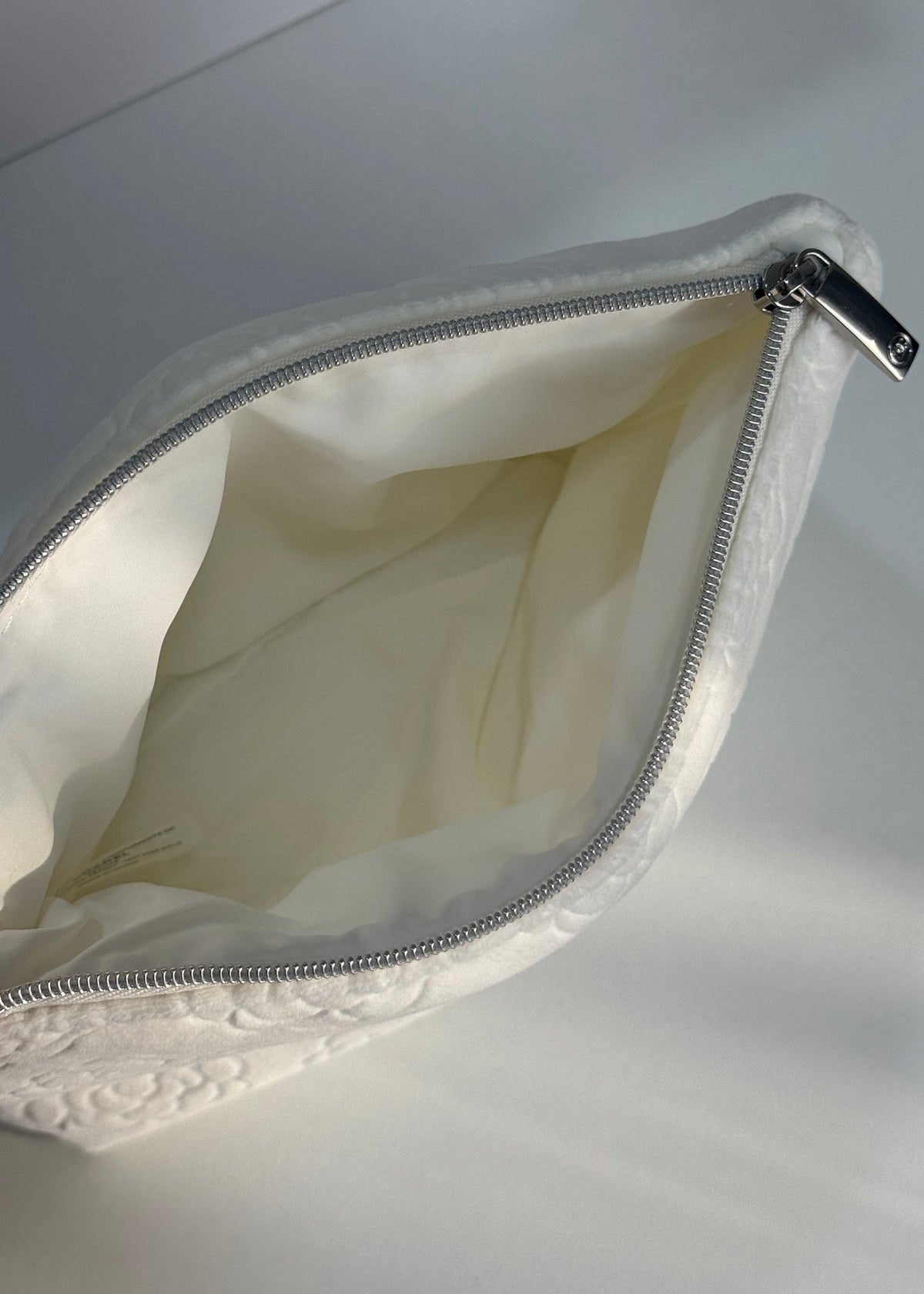 An open Chanel beauty pouch with a textured white exterior and cream-colored interior lining, shown with a white Chanel box in the background.