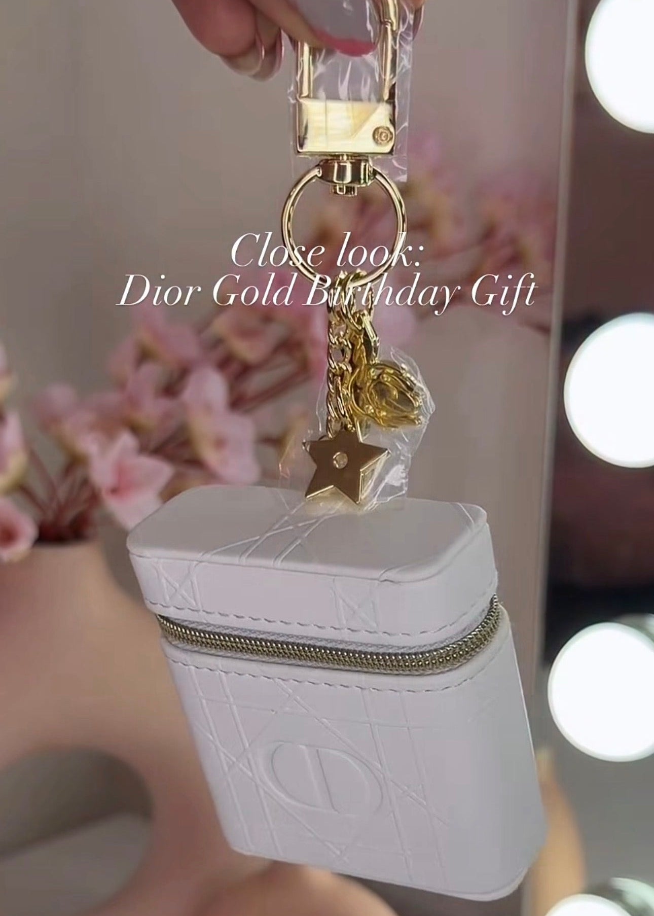 A close-up of a hand holding a white Dior-themed zippered case with gold hardware and charms, including a star charm and a clear lock. The background shows a softly blurred vanity mirror with light bulbs and pink flowers.