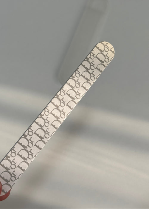 A close-up of a silver nail file featuring a repeating Dior Oblique monogram pattern, held diagonally against a soft-focus light gray background.