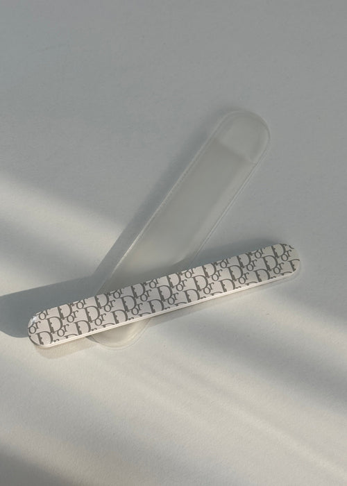 A nail file with a white and black Dior Oblique monogram pattern, resting on a translucent clear plastic nail file holder against a light gray background with soft diagonal shadows.