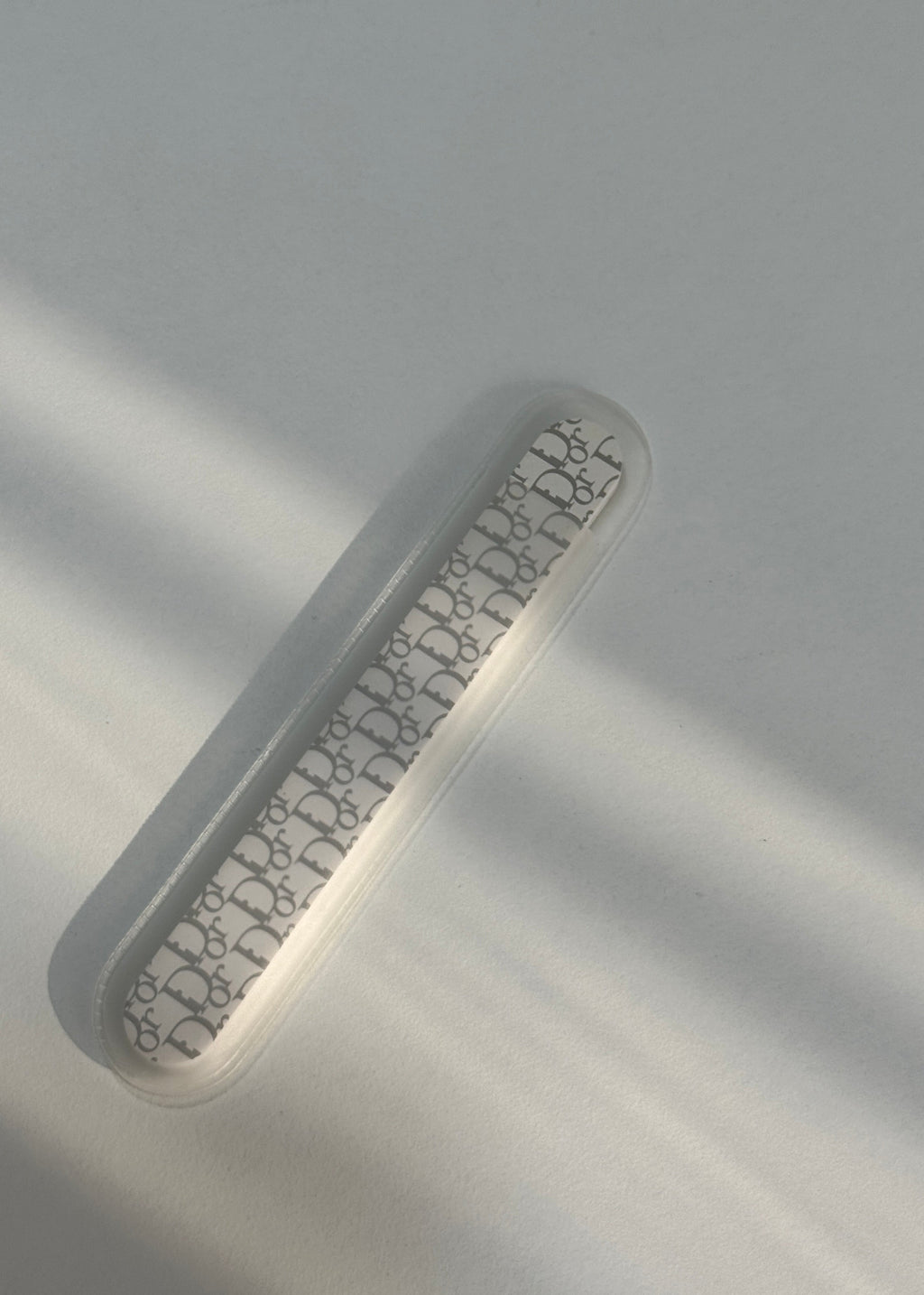 A clear, elongated plastic case with a repeating black Dior Oblique monogram pattern, shown on a light gray background with soft diagonal shadows.