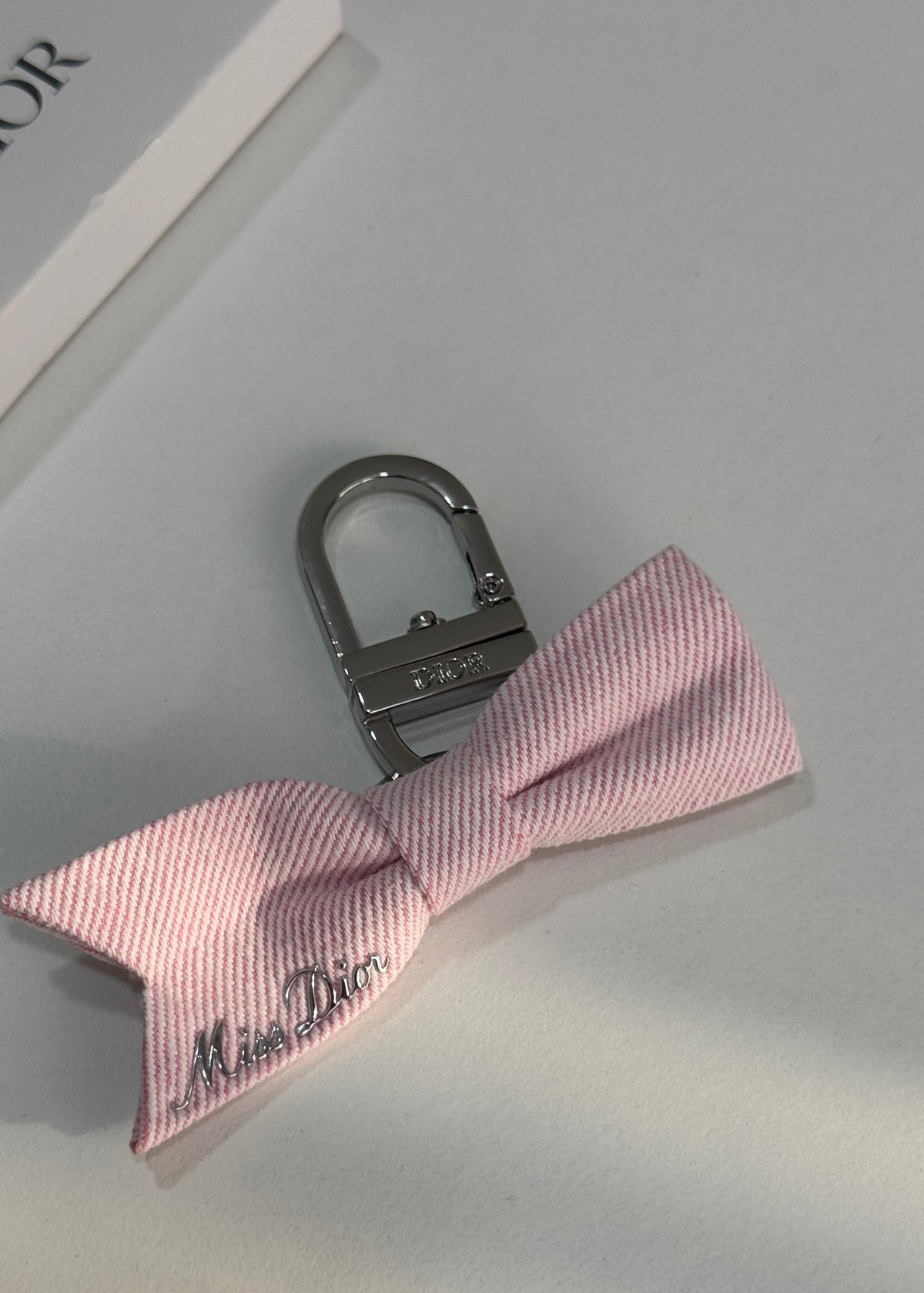 A pink striped bow tie keychain with a silver-tone metal clasp, featuring the 'Miss Dior' logo embroidered on the ribbon, displayed on a light gray surface.