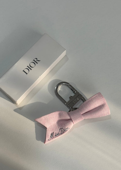A pink striped bow tie keychain with a silver metal clasp, displayed next to its white Dior box on a light gray surface.