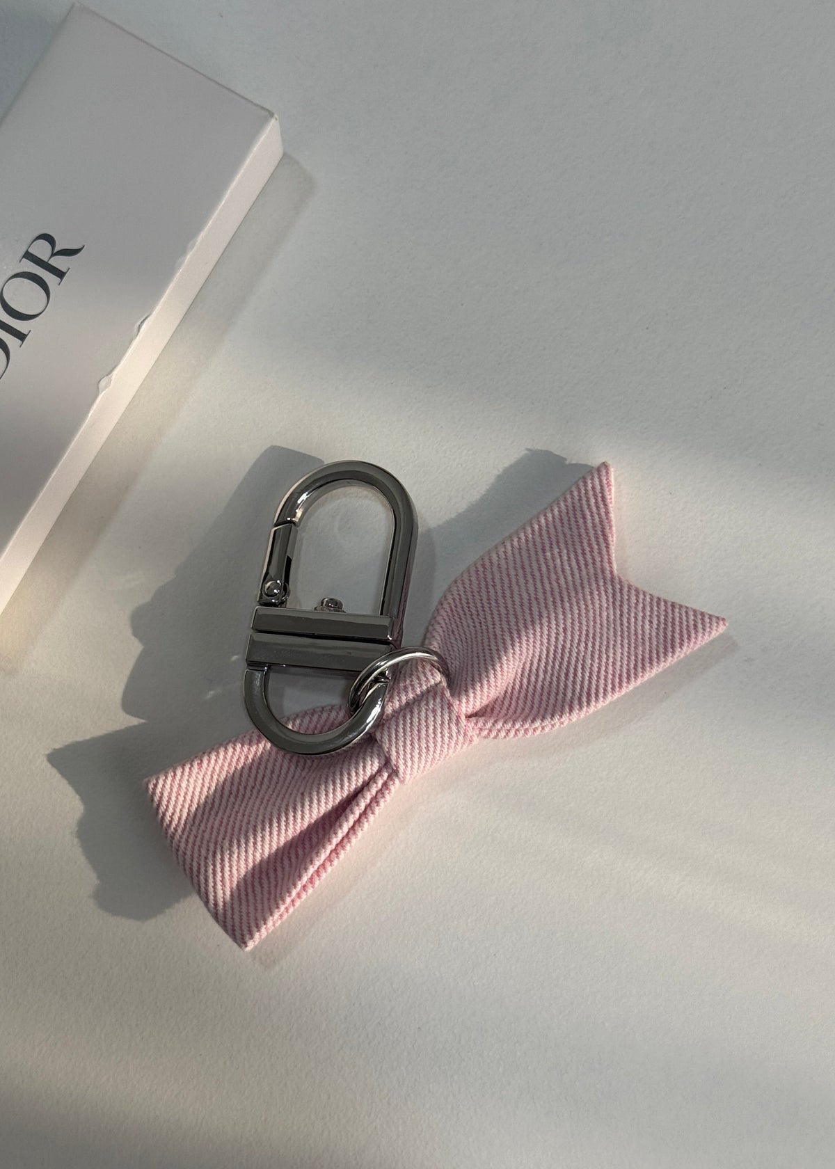 A pink striped bow keychain with a silver metal clasp, displayed on a light gray surface next to a white Dior box.