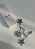 A pair of silver-tone Dior star and chain earrings with a square pendant charm, displayed next to a white Dior box on a light gray surface.