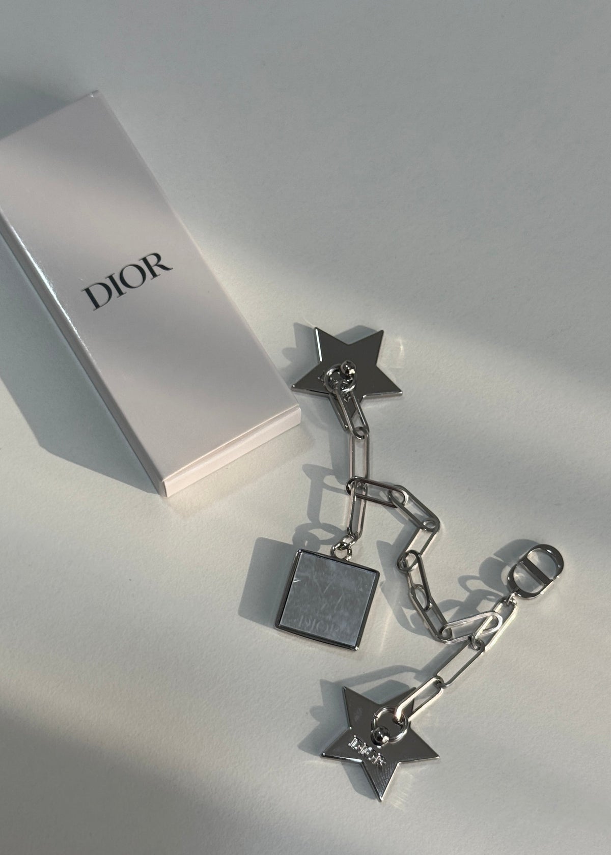 A pair of silver-tone Dior star and chain earrings with a square pendant charm, displayed next to a white Dior box on a light gray surface.