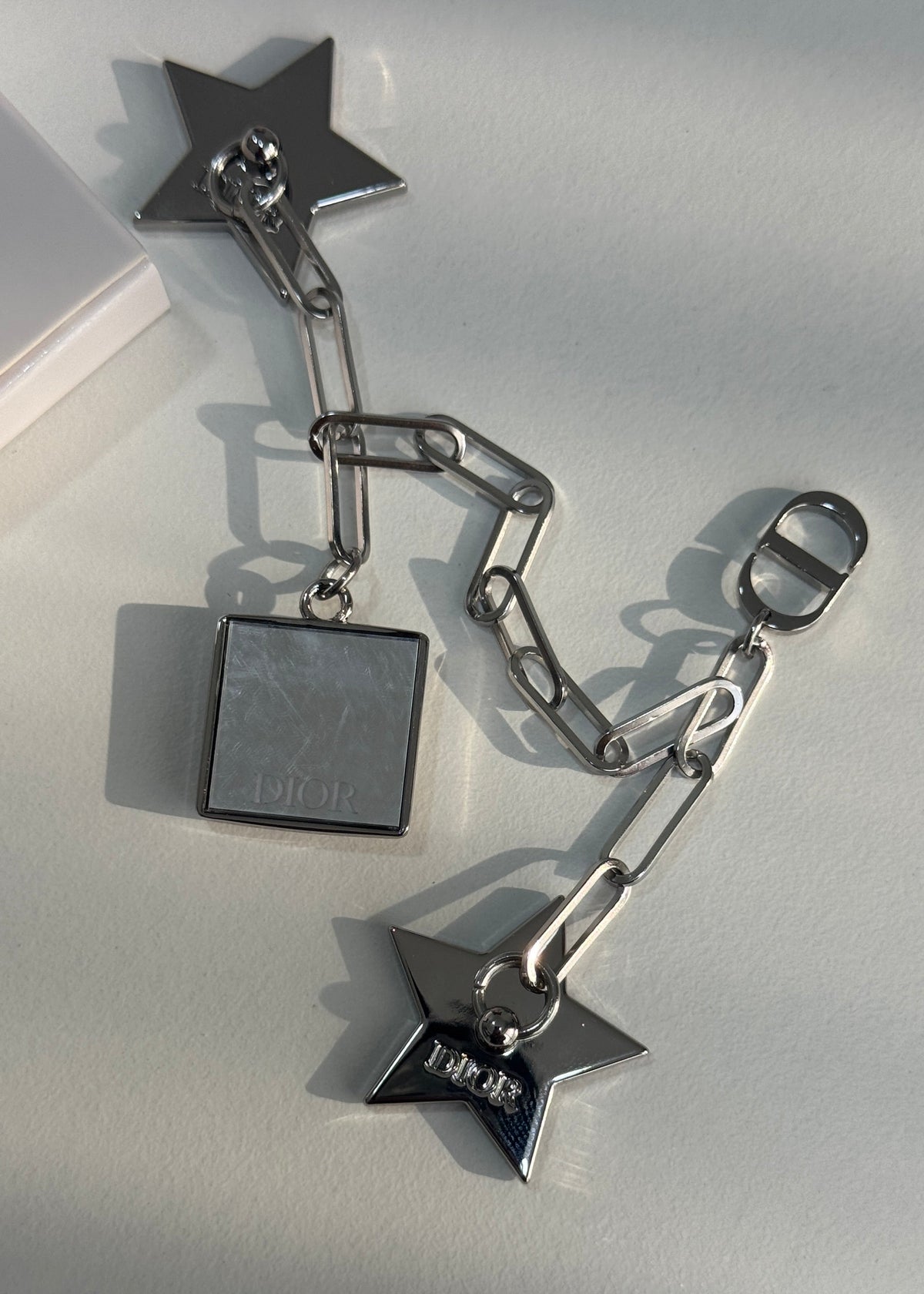 A silver-toned metal accessory set featuring a square pendant, two star-shaped charms, and a chain with a lobster clasp, photographed on a light surface with strong sunlight and shadows.