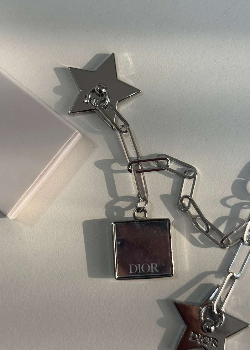 A silver-tone Dior star and chain accessory with a square Dior box charm, photographed on a light neutral surface with a white box edge visible.