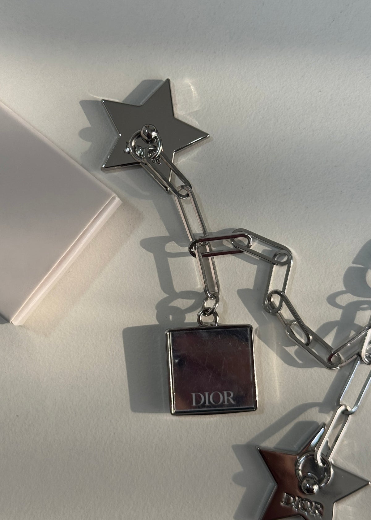A silver-tone Dior star and chain accessory with a square Dior box charm, photographed on a light neutral surface with a white box edge visible.
