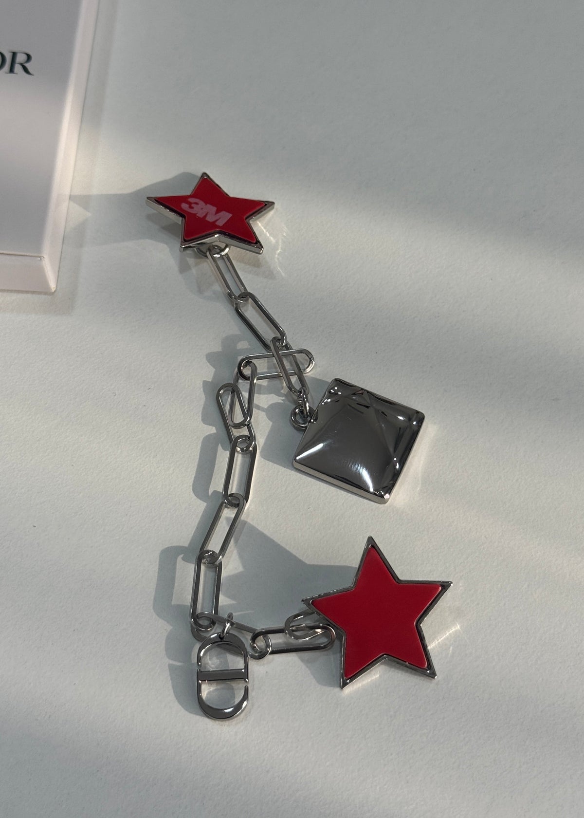 A silver-tone chain accessory with two red star charms and a small metallic square charm, photographed on a light gray surface with part of a white box visible at the upper left.
