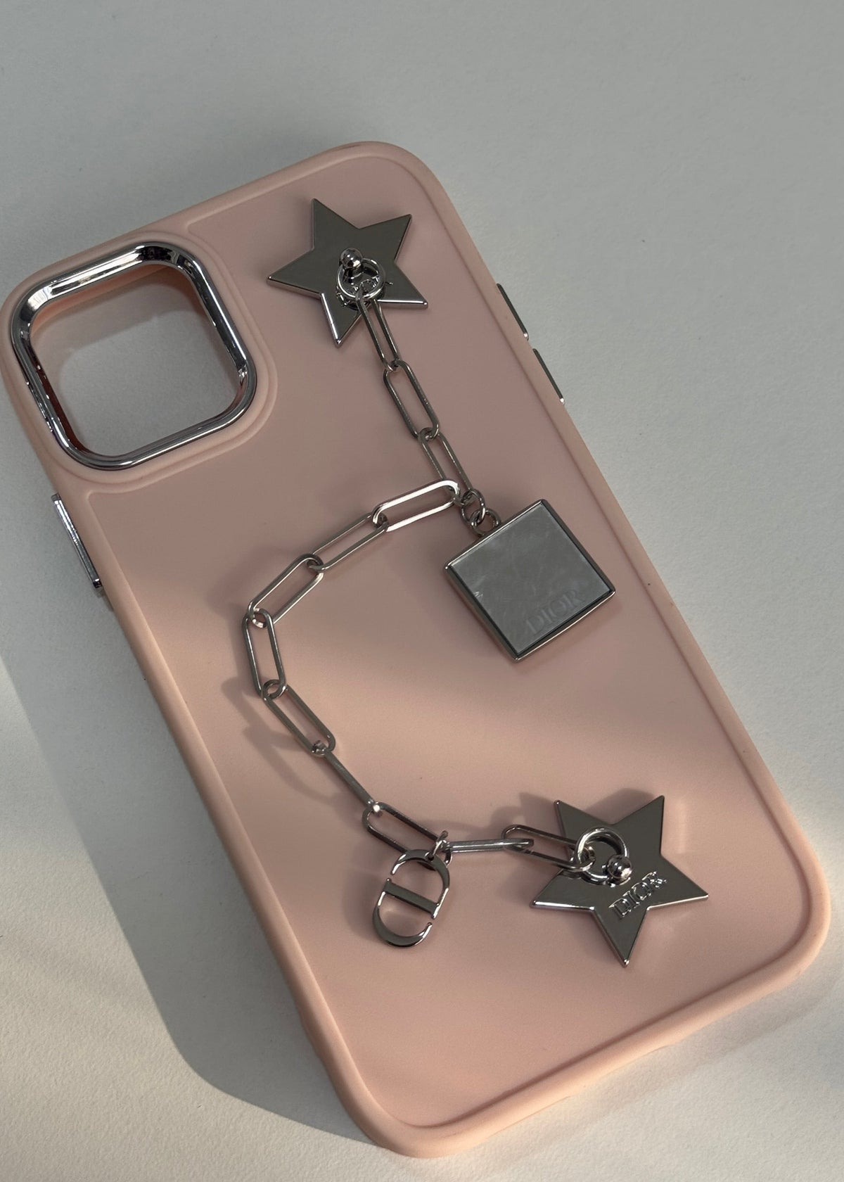 A light pink phone case with a silver metal chain accessory featuring star and square charms, displayed on a neutral gray background.