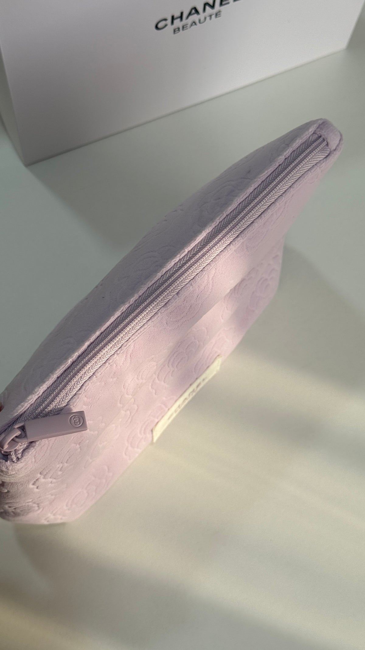 A light purple Chanel beauty pouch with a textured fabric and a matching zipper pull, shown next to its white Chanel box.
