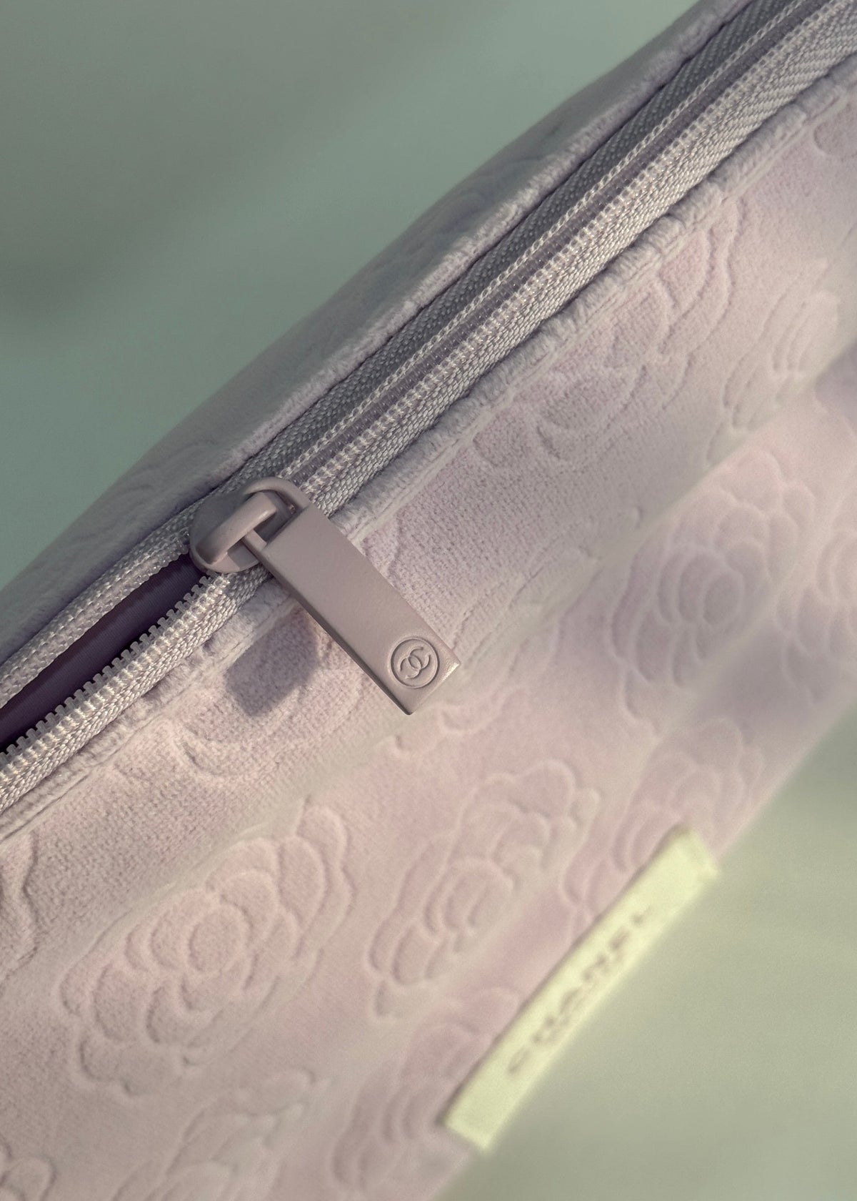 A close-up of a light pink or pale lavender zippered pouch with an embossed floral pattern, featuring a silver zipper pull and a small cream-colored brand tag.