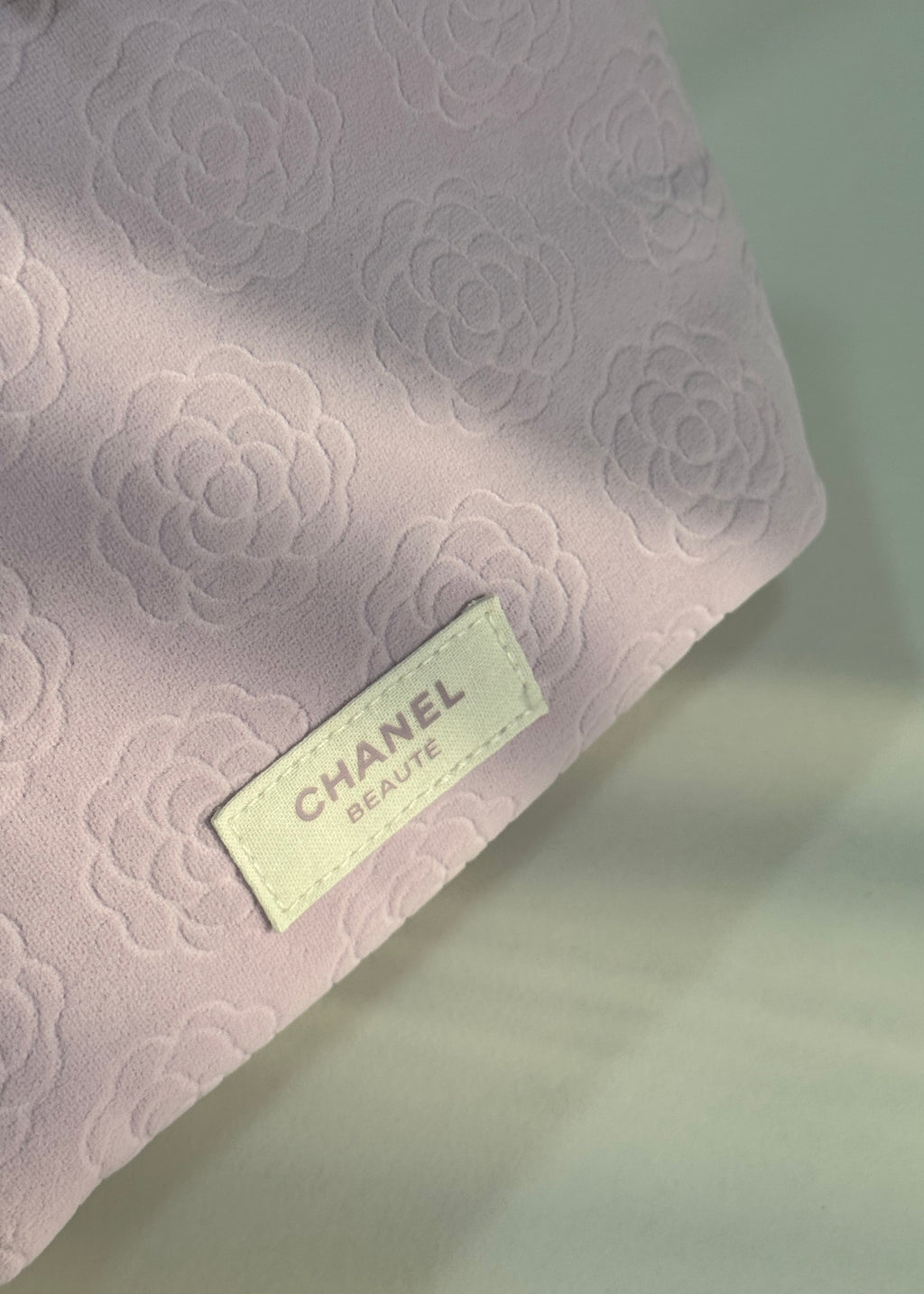 A close-up of a light pink Chanel Beauty pouch with an embossed floral pattern and a cream-colored brand label.