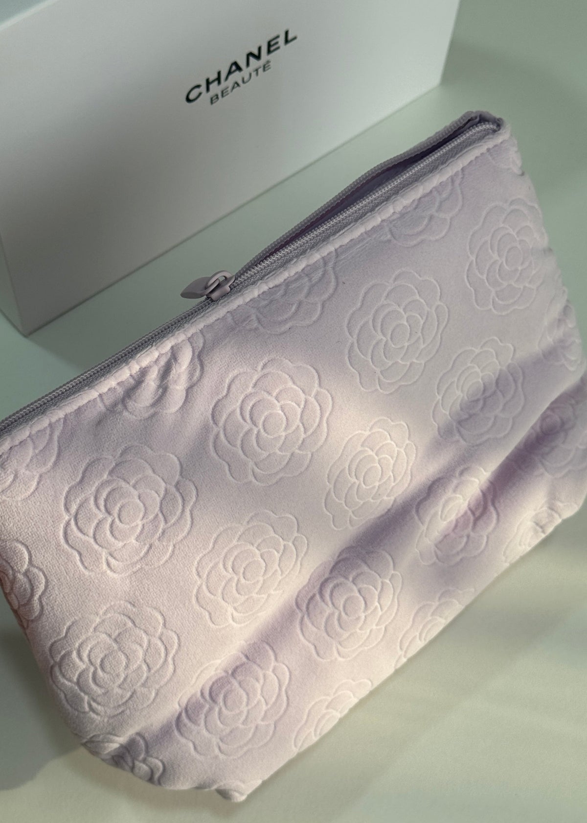A light lavender Chanel cosmetic pouch with an embossed floral camellia pattern, shown next to its white Chanel Beaute box.