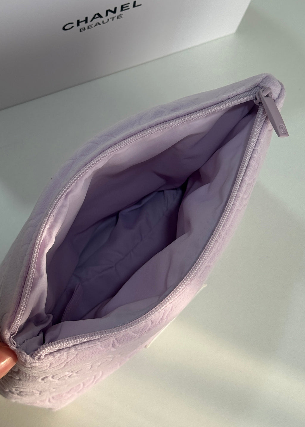A light purple Chanel cosmetic pouch shown open, with its interior visible and a Chanel box in the background.