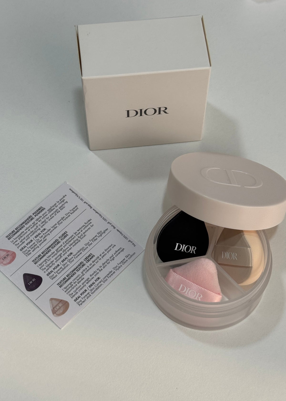 Dior powder foundation compact with a beige lid, shown open next to its cream-colored Dior box and a small instruction card on a light gray surface.