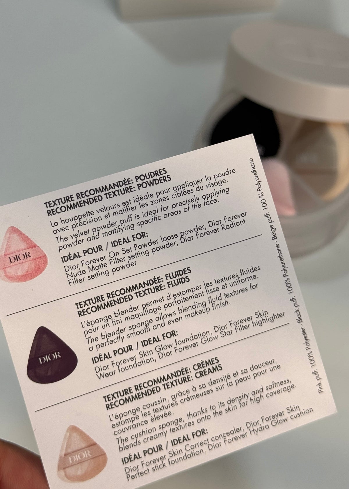 A hand holds a printed card showing product information and shade swatches for a powder foundation, with a blurred makeup palette and white box in the background.