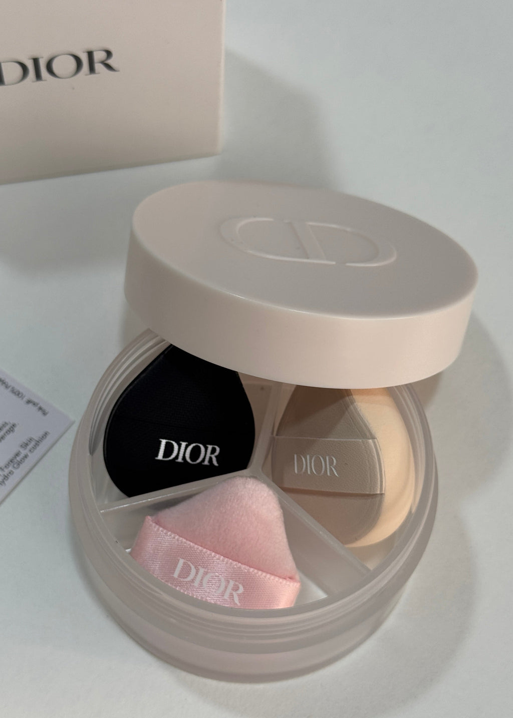 Dior makeup compact with a light beige lid and a clear round base showing three makeup sponges: one black, one pink, and one beige, with Dior branding visible on the sponges and compact.
