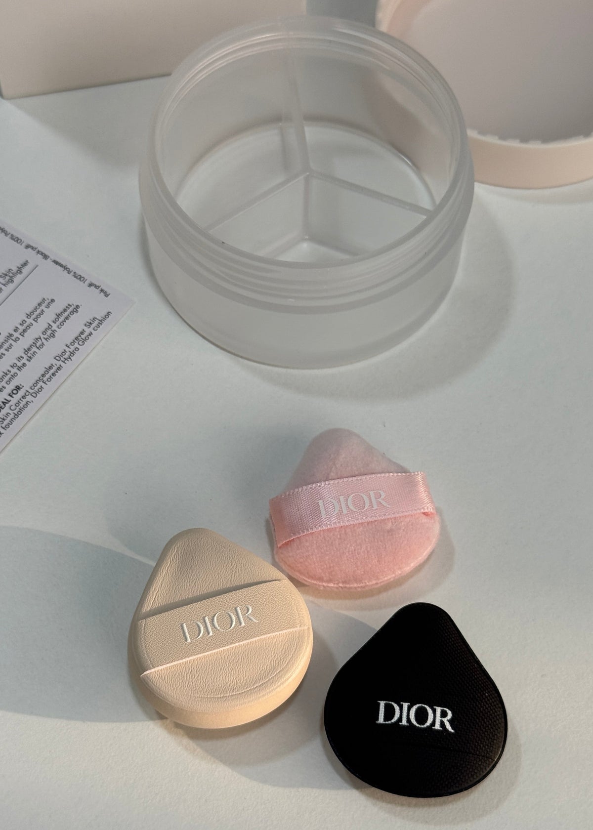 Dior makeup applicator sponges and compact case on a clean white surface, including a beige teardrop-shaped sponge with a beige cap, a pink teardrop-shaped sponge with a pink cap, and a clear round multi-compartment container.