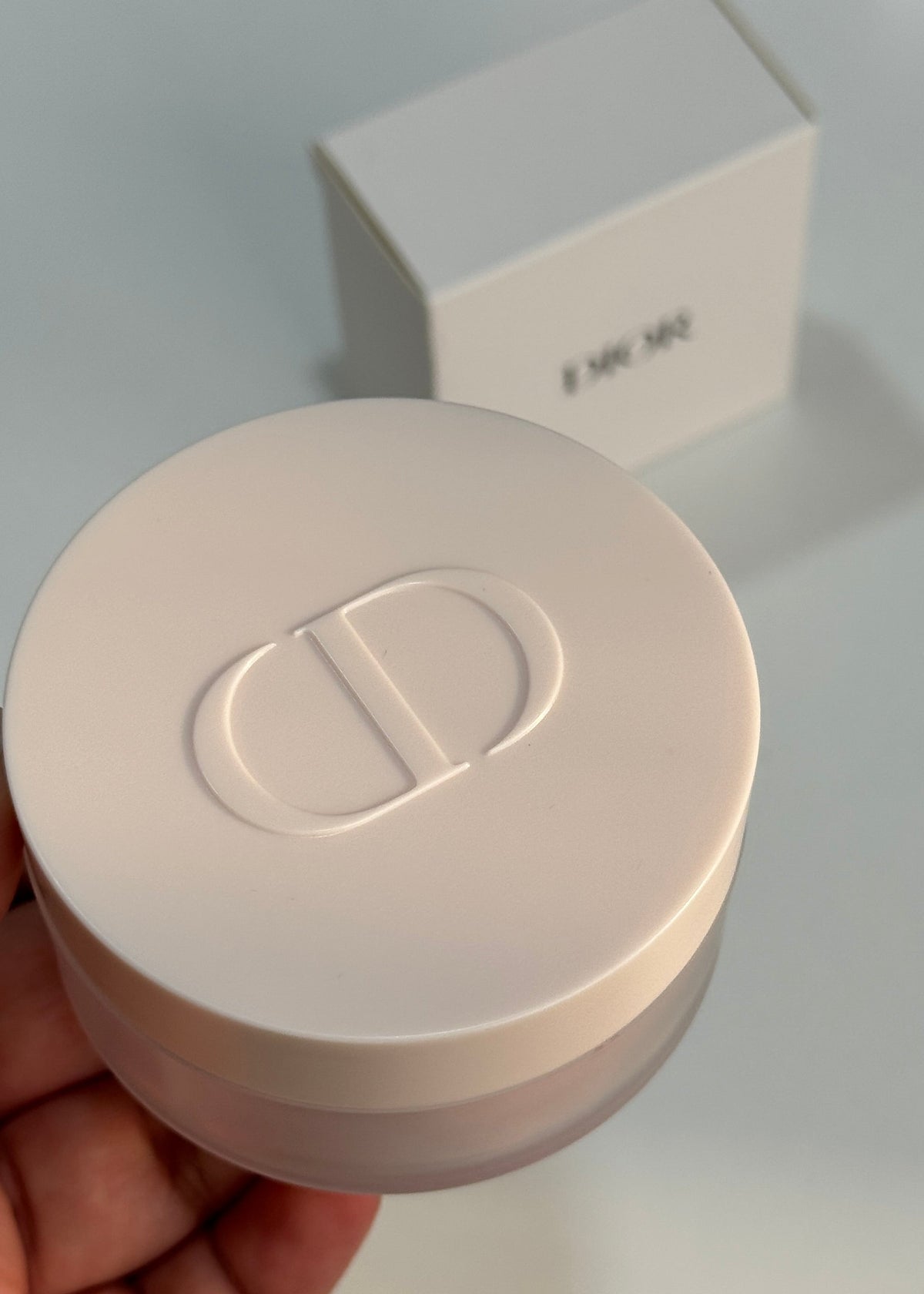 A hand holding a round, off-white cosmetic jar with a debossed logo on the lid, with a matching off-white box in the background.