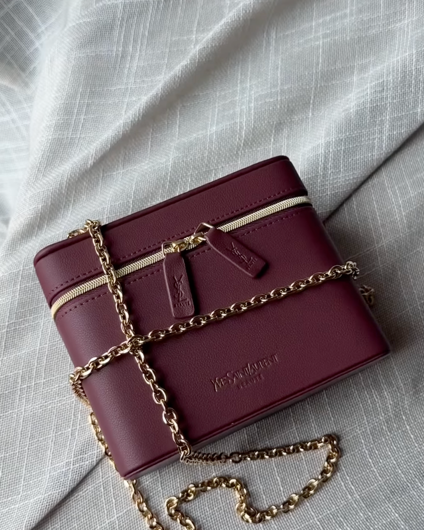 A burgundy leather crossbody bag with a gold chain strap, laid flat on a textured light gray fabric background. The brand name 'Burberry' is embossed on the front.