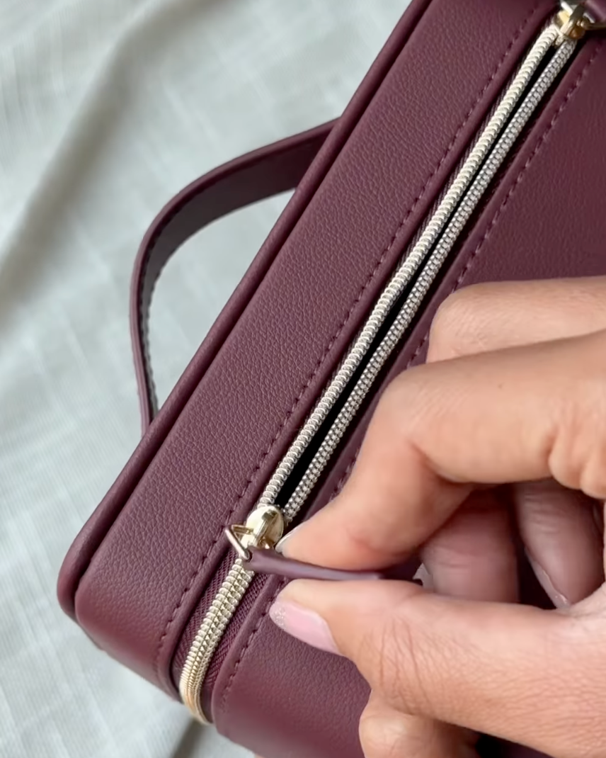 A close-up of a burgundy pebbled leather zippered case being opened by a hand, showing a gold-tone zipper with a pull tab.