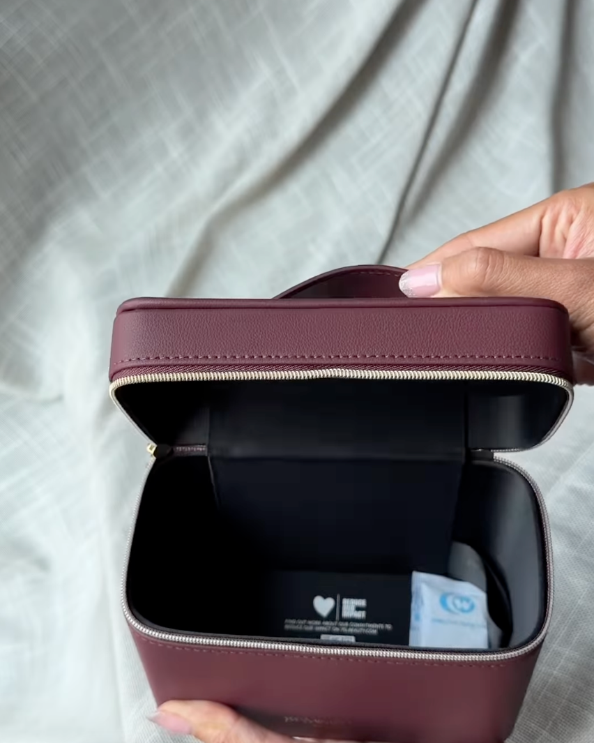 A person holds open a small rectangular zippered case in a dusty burgundy or mauve-brown color, showing a black interior with a small card and a white item inside. The background is a light gray fabric backdrop.