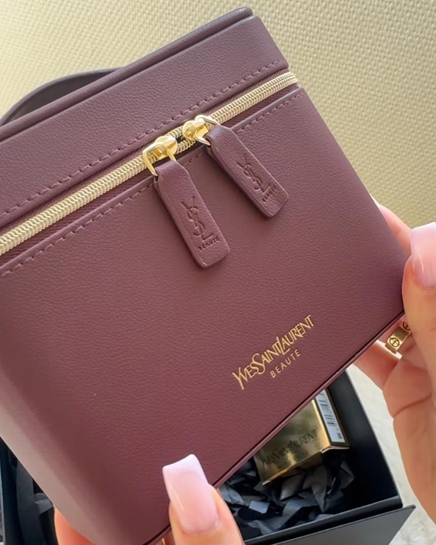 A close-up of a person holding a Saint Laurent cosmetic bag in a dusty rose color with gold hardware and logo. The Saint Laurent logo is clearly visible on the front of the bag.
