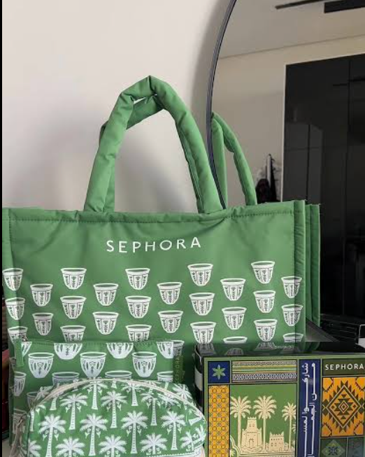 A green Sephora tote bag with white cup and palm tree patterns, displayed next to a matching zippered pouch and a Sephora gift box on a glass shelf.